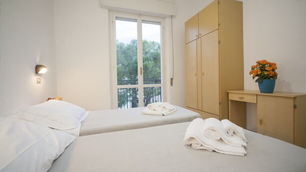 A bedroom with two beds, a wooden wardrobe, a window with a view, and a small table with a vase of flowers. - Blue Sky (Rimini)
