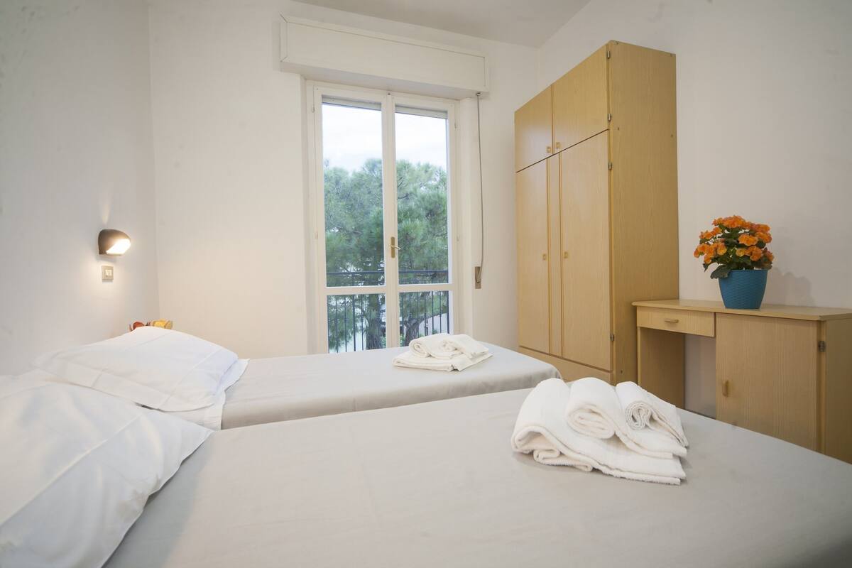 A bedroom with two beds, a wooden wardrobe, a window with a view, and a small table with a vase of flowers.