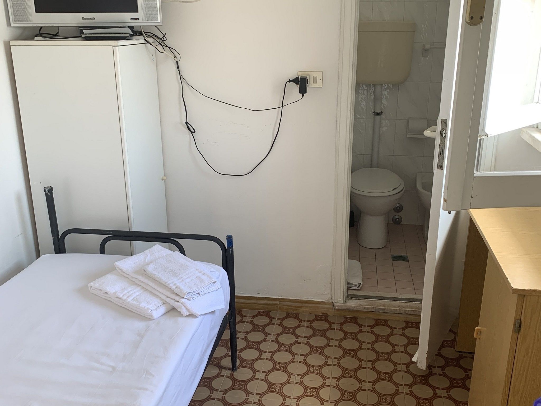A small, simple room with a bed, a television on a cabinet, and a bathroom visible through an open door.