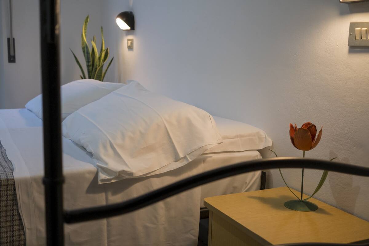 A neatly made bed with white linens, a bedside table with a vase of a single flower, and a wall-mounted light fixture.