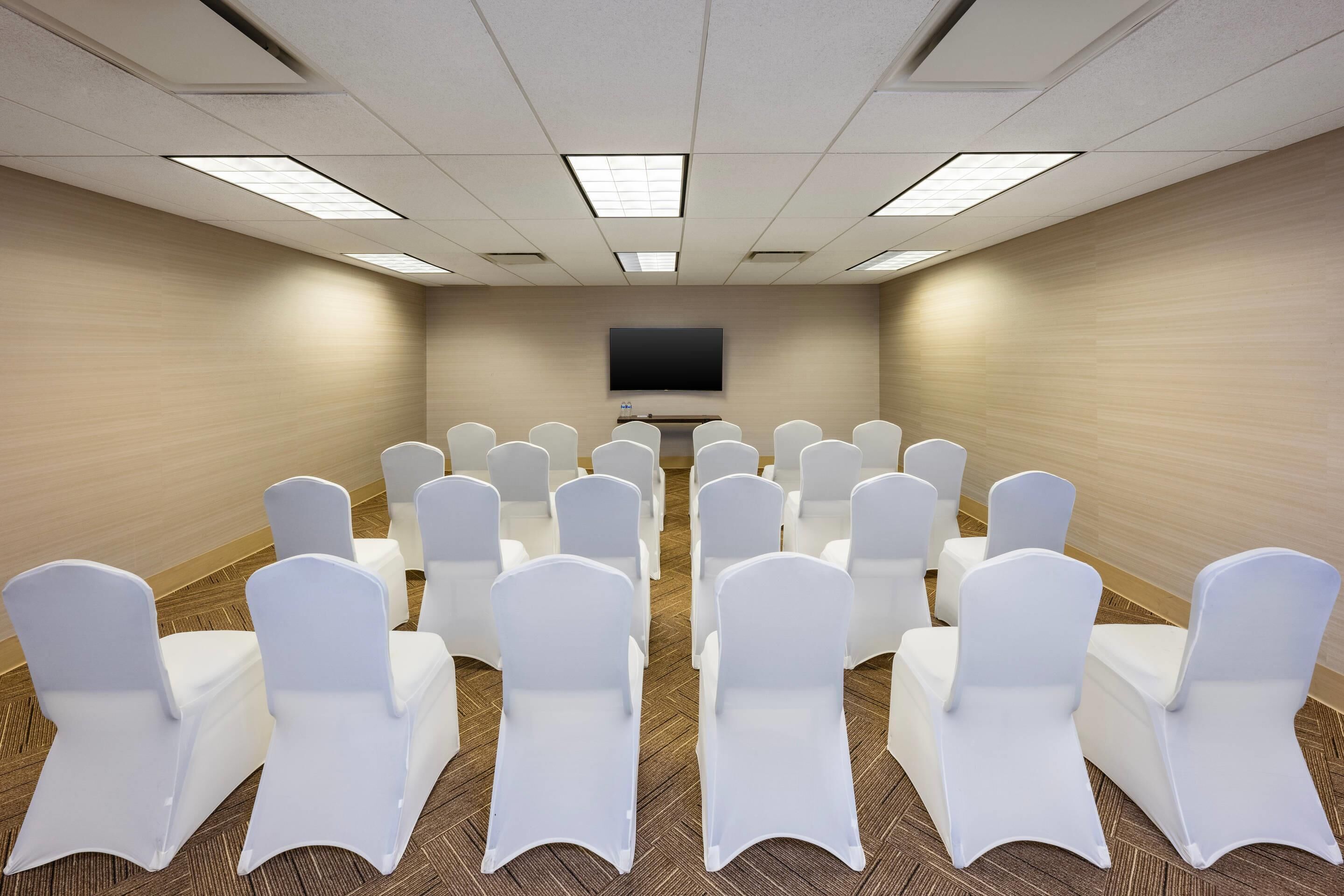 A conference room with rows of white chairs, a flat-screen TV on the wall, and carpeted flooring.