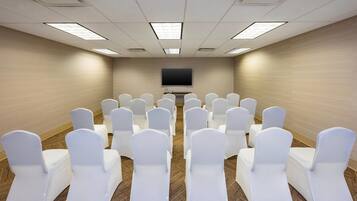 A conference room with rows of white chairs, a flat-screen TV on the wall, and carpeted flooring.