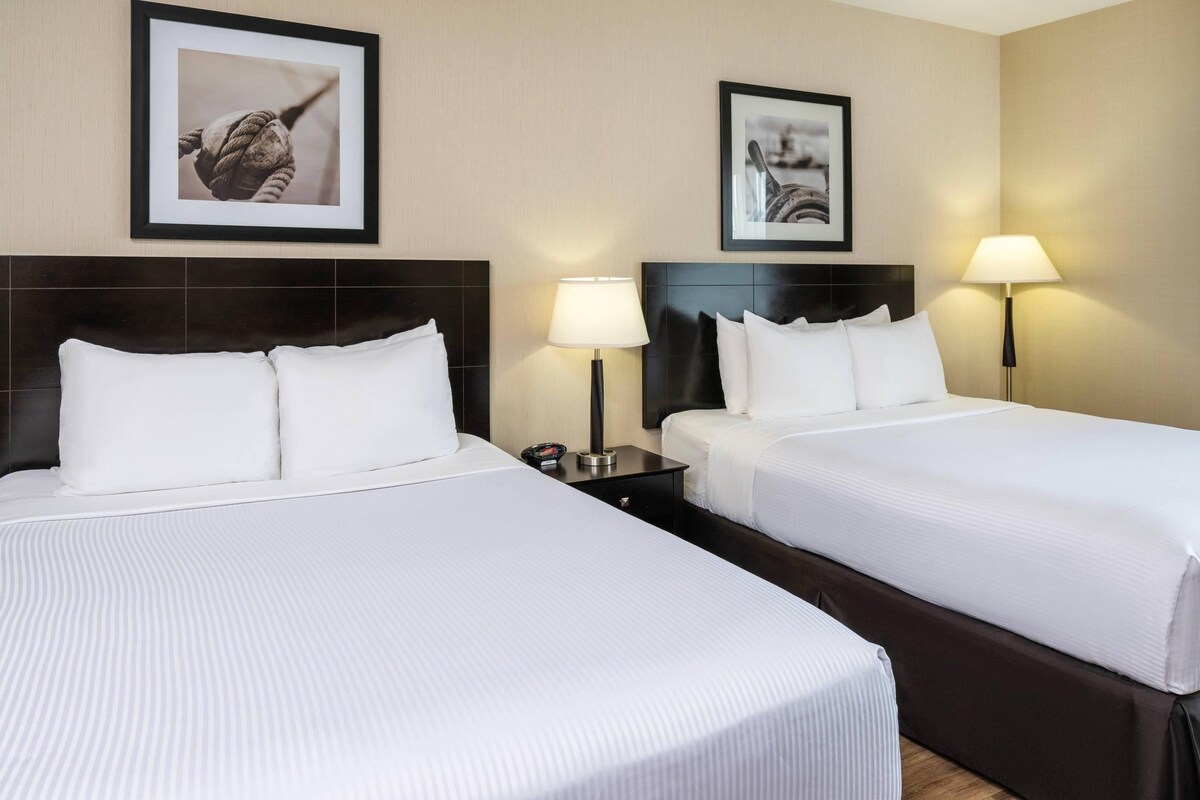 A hotel room with two beds, a headboard, bedside tables, lamps, and framed pictures on the wall.