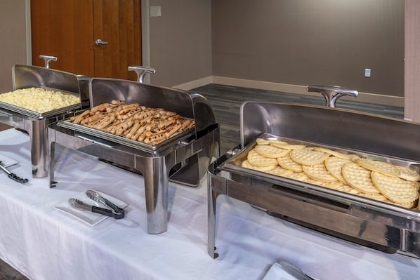 Una mesa de buffet con diferentes alimentos en recipientes metálicos, una puerta al fondo y un suelo alfombrado.