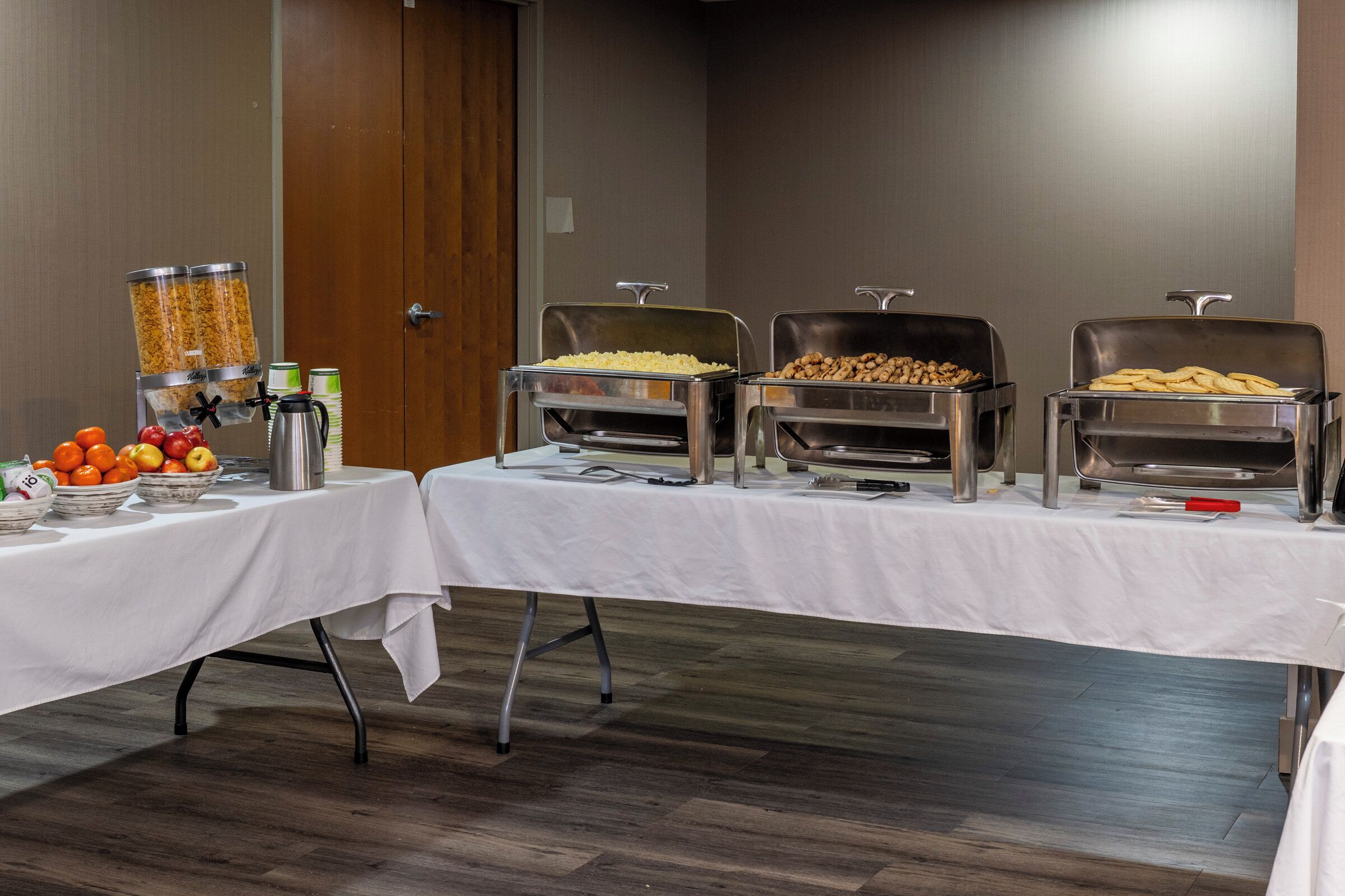 A buffet setup with various food containers, a fruit bowl, and a coffee dispenser on a table.