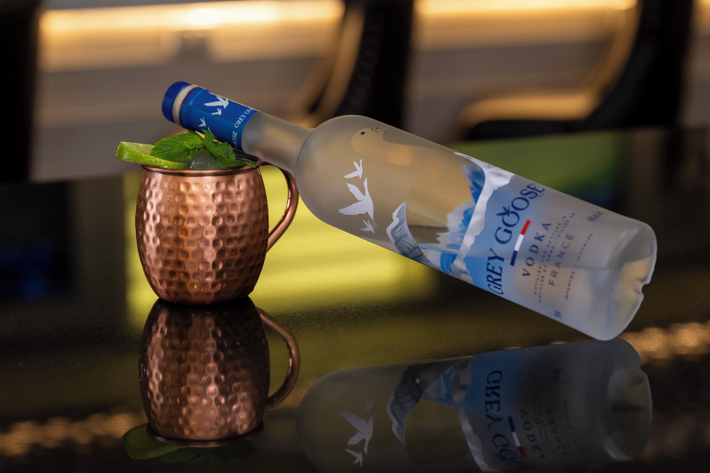 A bottle of Grey Goose vodka with a bird design, resting on a copper mug with mint leaves.