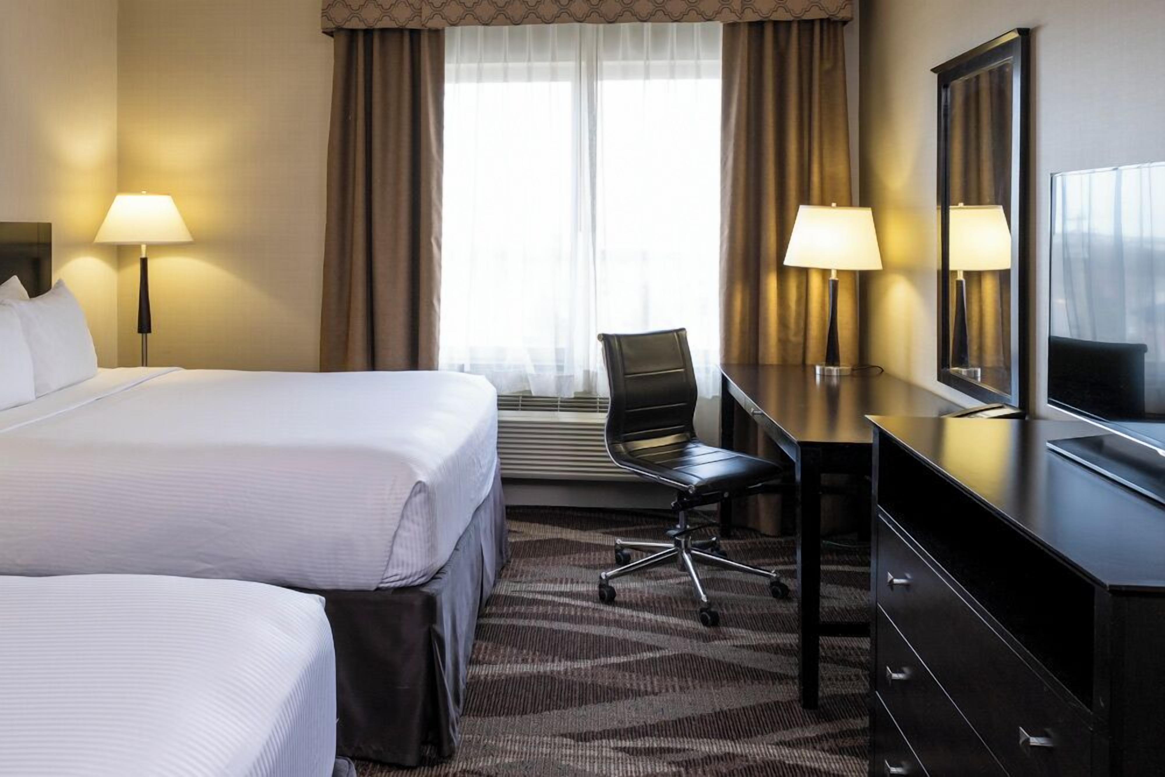 A hotel room with a large bed, two bedside lamps, a desk with a chair, a mirror, and a window with curtains.