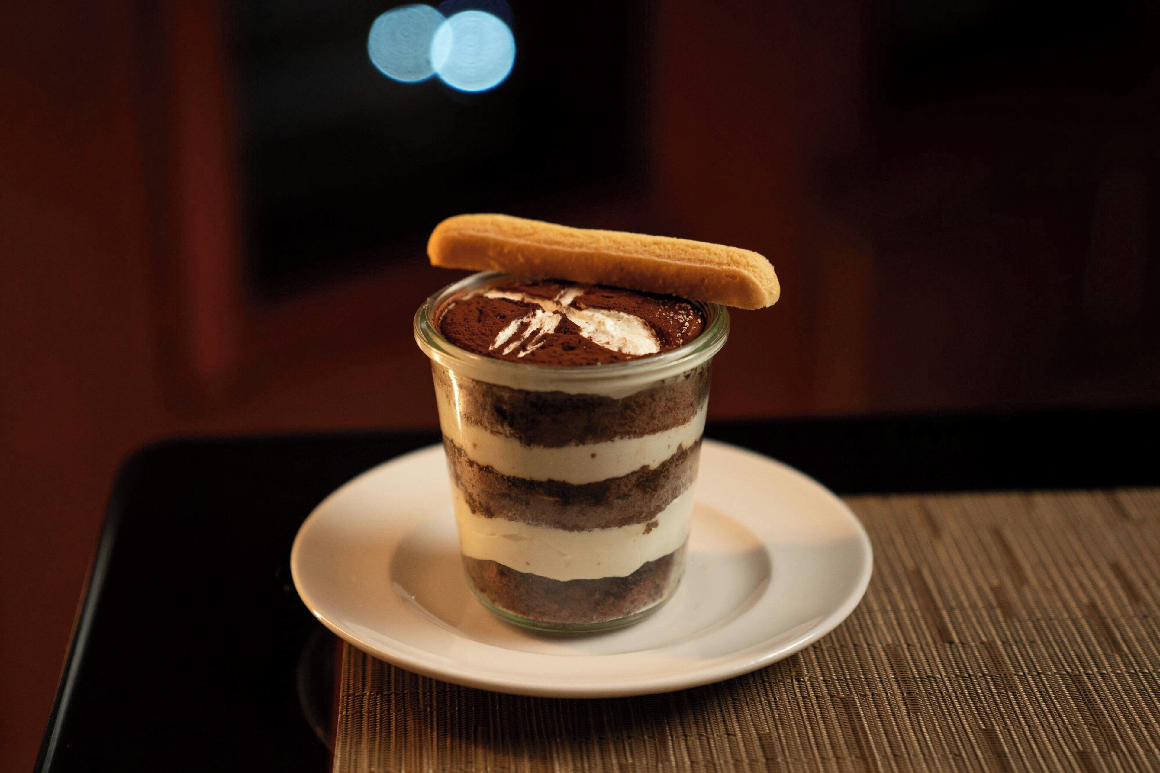 Een gelaagd dessert in een glazen pot met een koekje erop, geplaatst op een wit bord op een tafel.