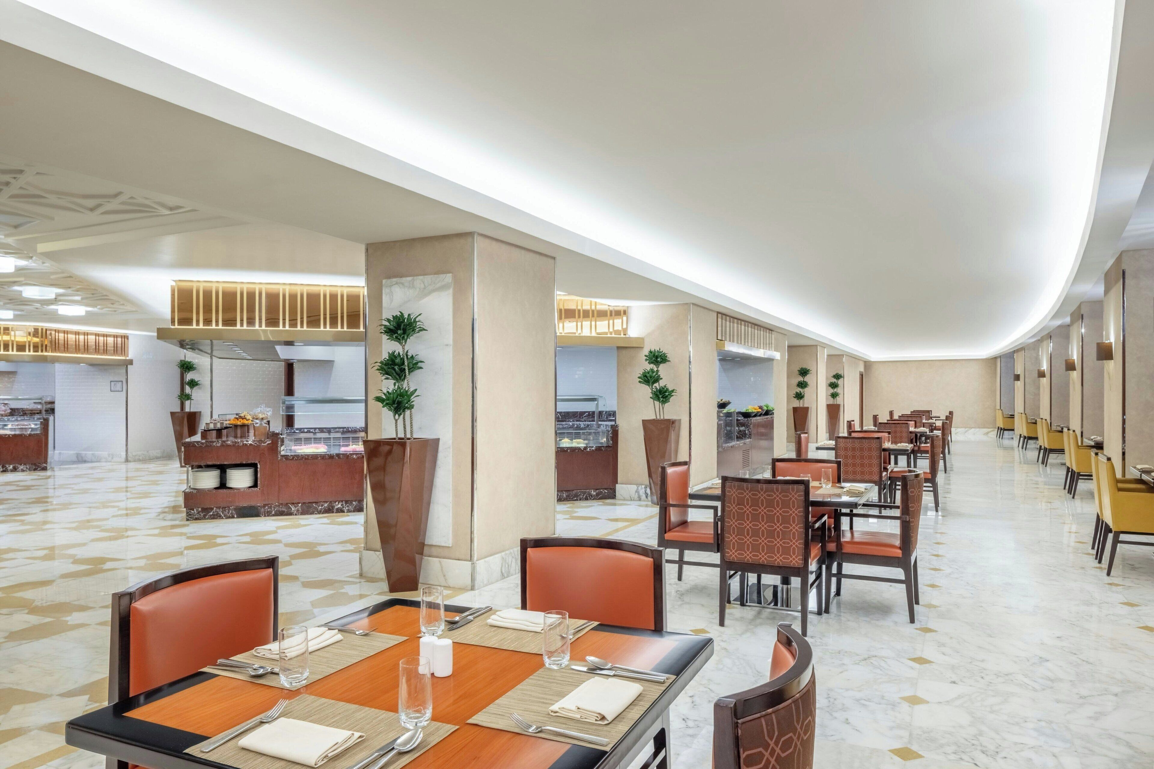 A spacious hotel dining area with multiple tables and chairs, a central counter with a display, and potted plants.