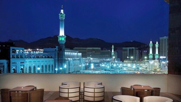 A rooftop lounge with wicker furniture and a view of a cityscape including a prominent mosque and skyscrapers.