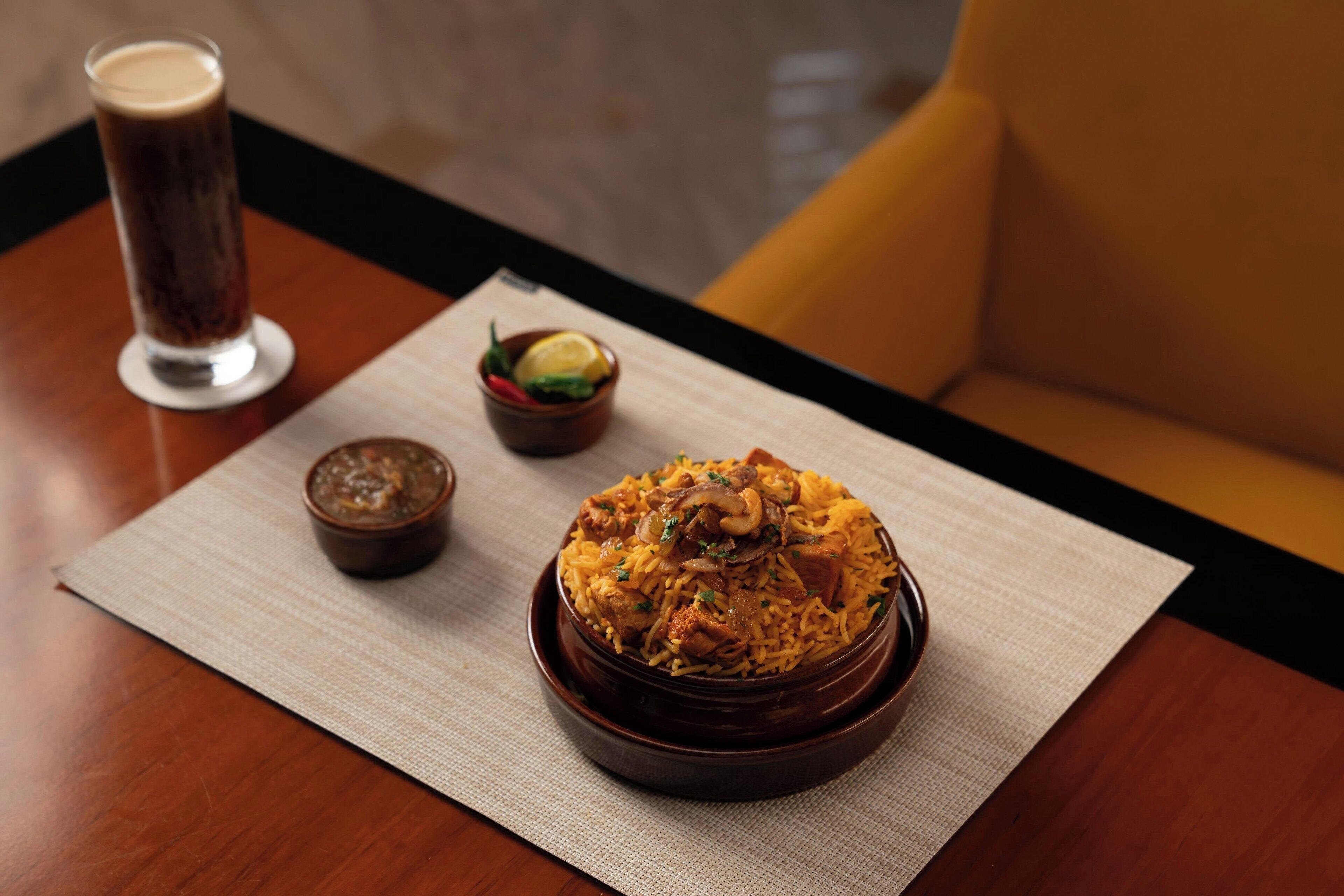 A table with a bowl of biryani, a small bowl of chutney, and a glass of beverage.