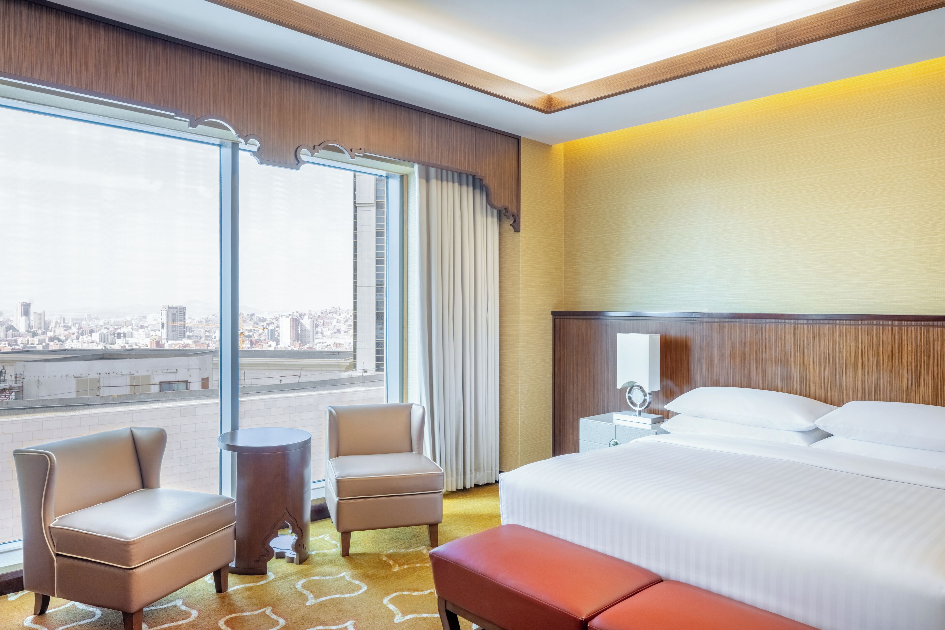 A hotel room with a large bed, two armchairs, a small table, and a view of the city through large windows.