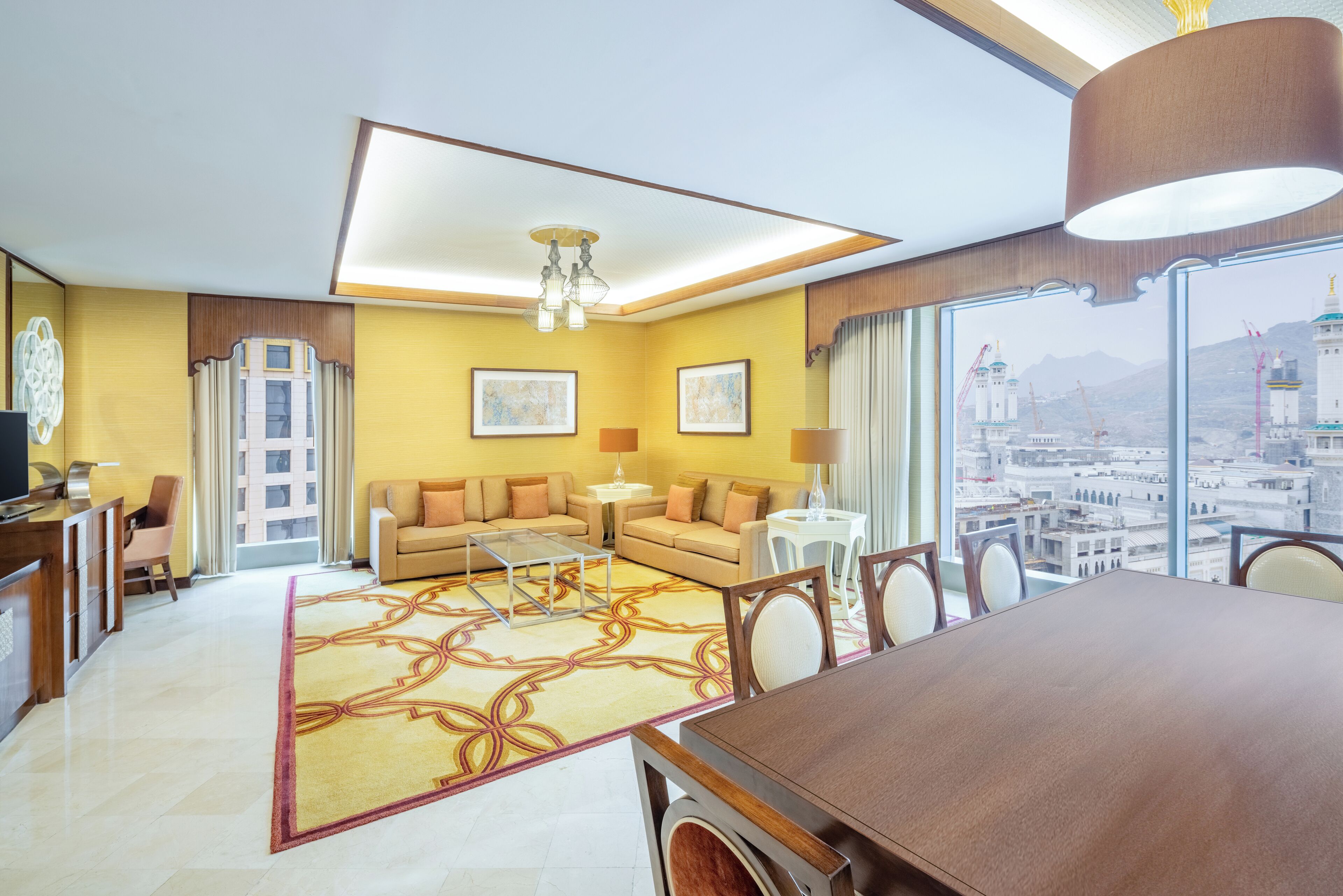 A modern hotel room with a large dining table, a sofa set, a coffee table, and a view of a cityscape through large windows.