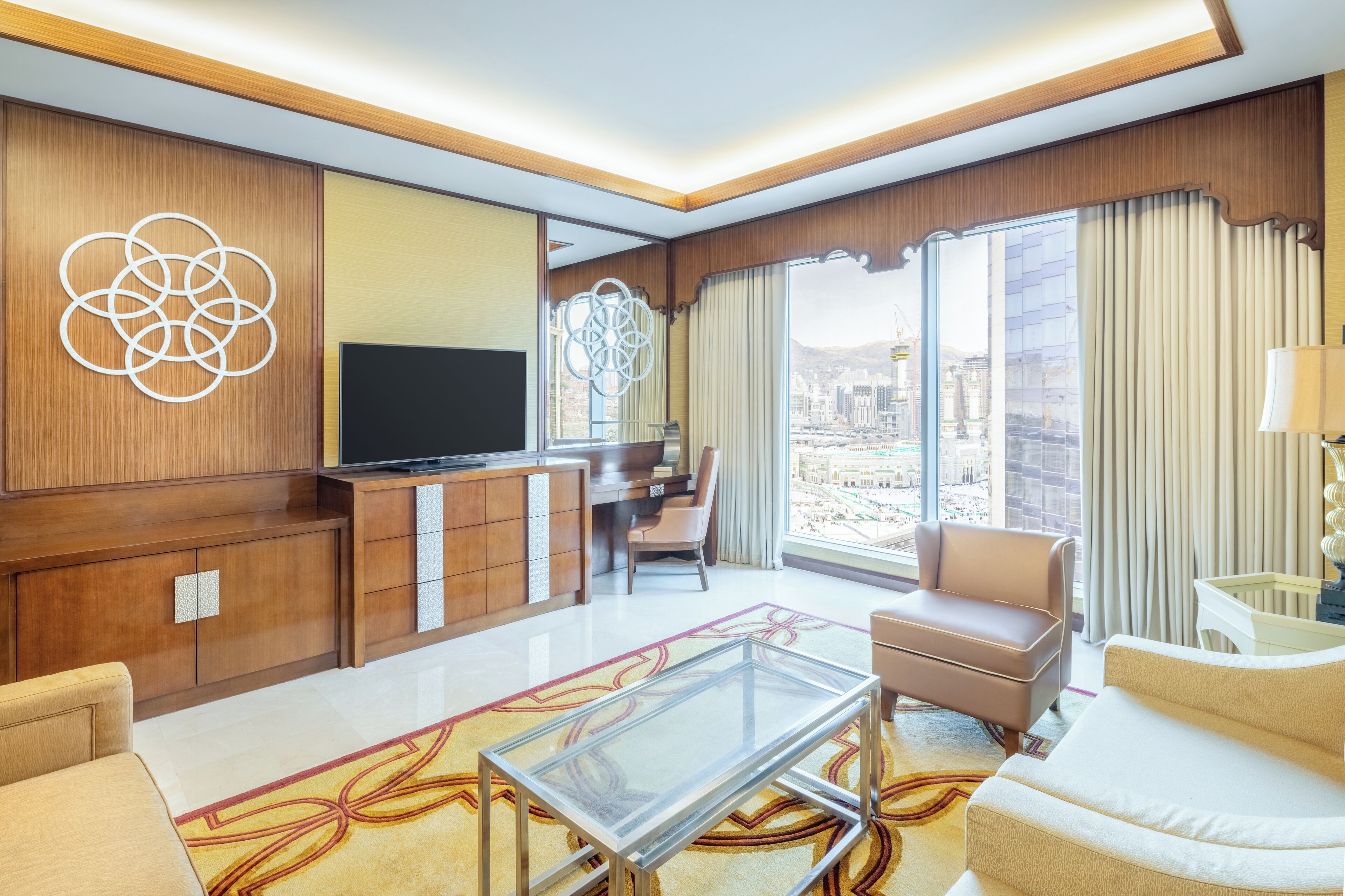 A modern hotel room with a large TV, comfortable seating, a glass coffee table, and a view of the city.