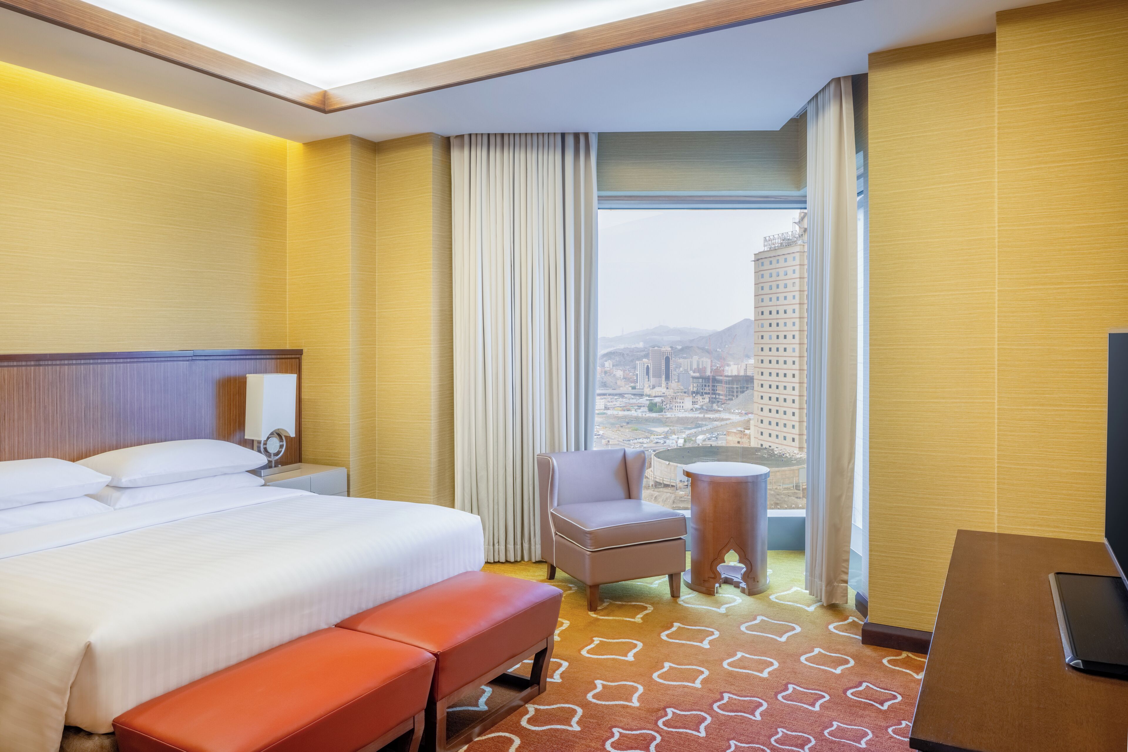 A hotel room with a large bed, a small table, a chair, and a view of a cityscape through a large window.