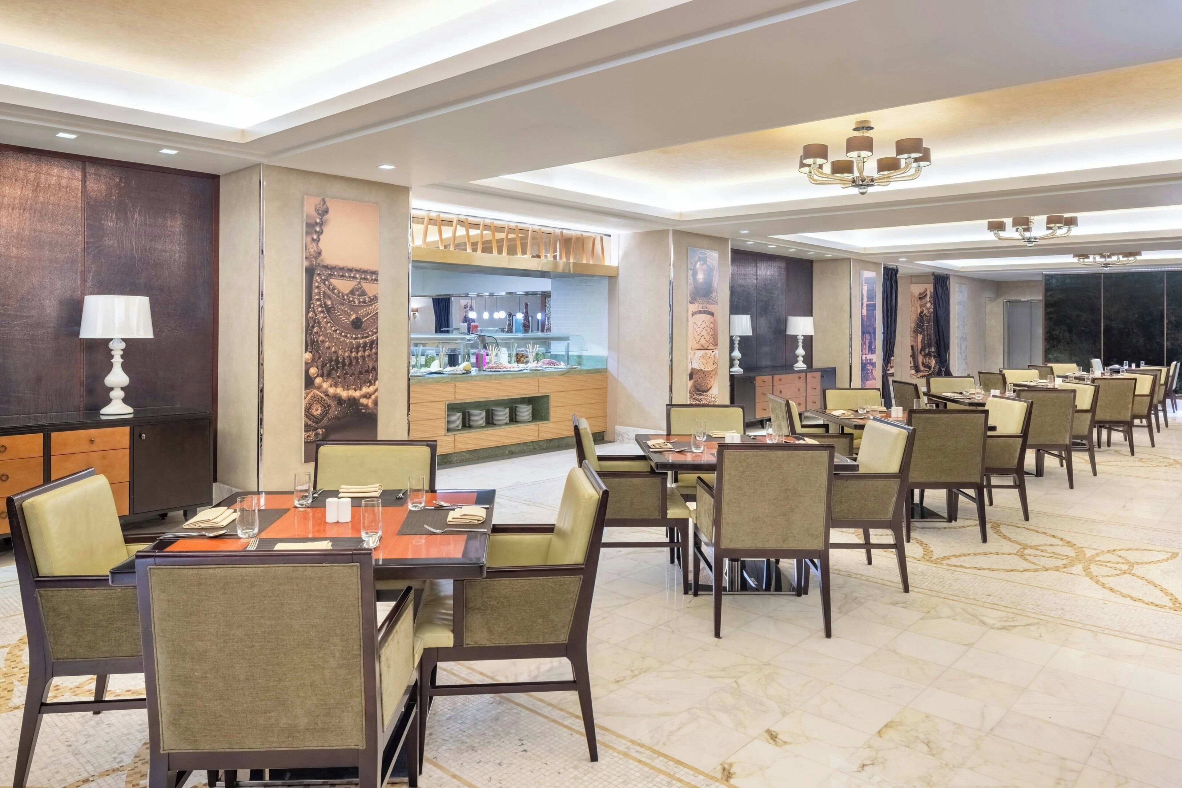 A hotel dining area with multiple tables and chairs, a bar area with glassware, and a large window providing a view.