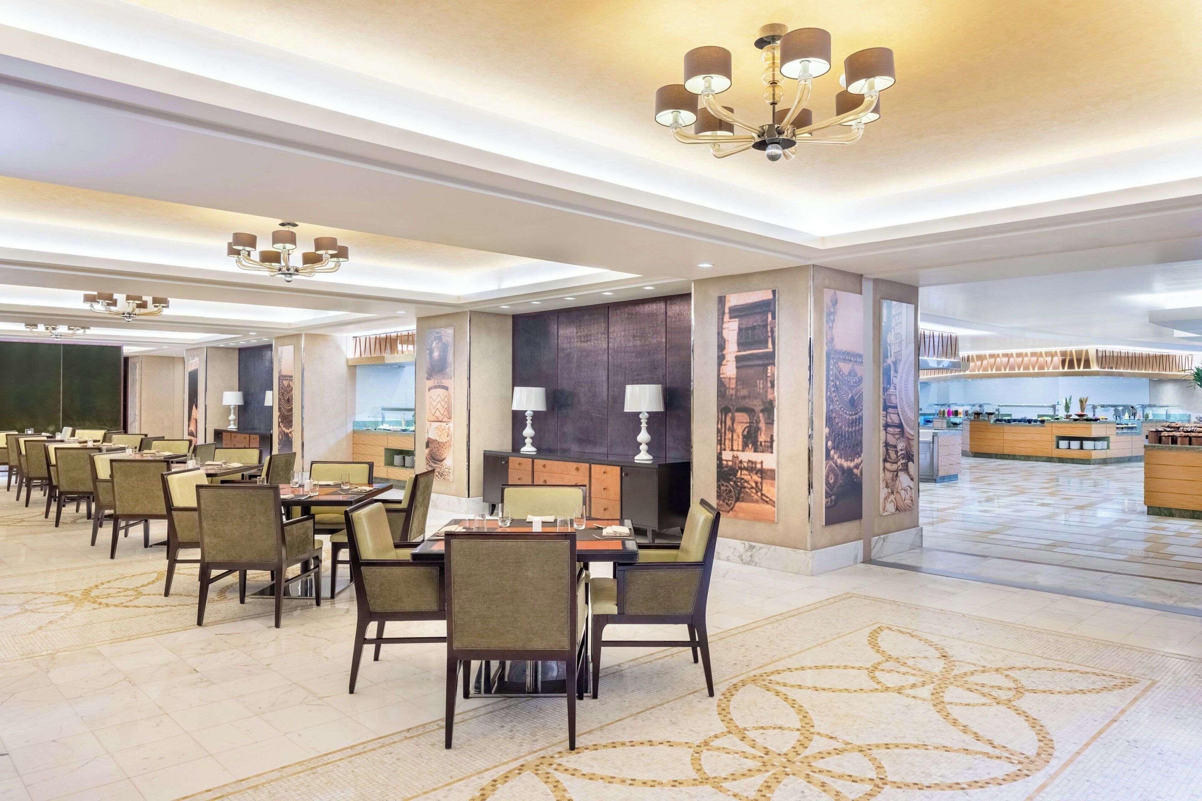 Une vaste salle à manger d’hôtel, dotée de nombreuses tables et chaises, d’un lustre central et offrant une vue sur la cuisine en arrière-plan.