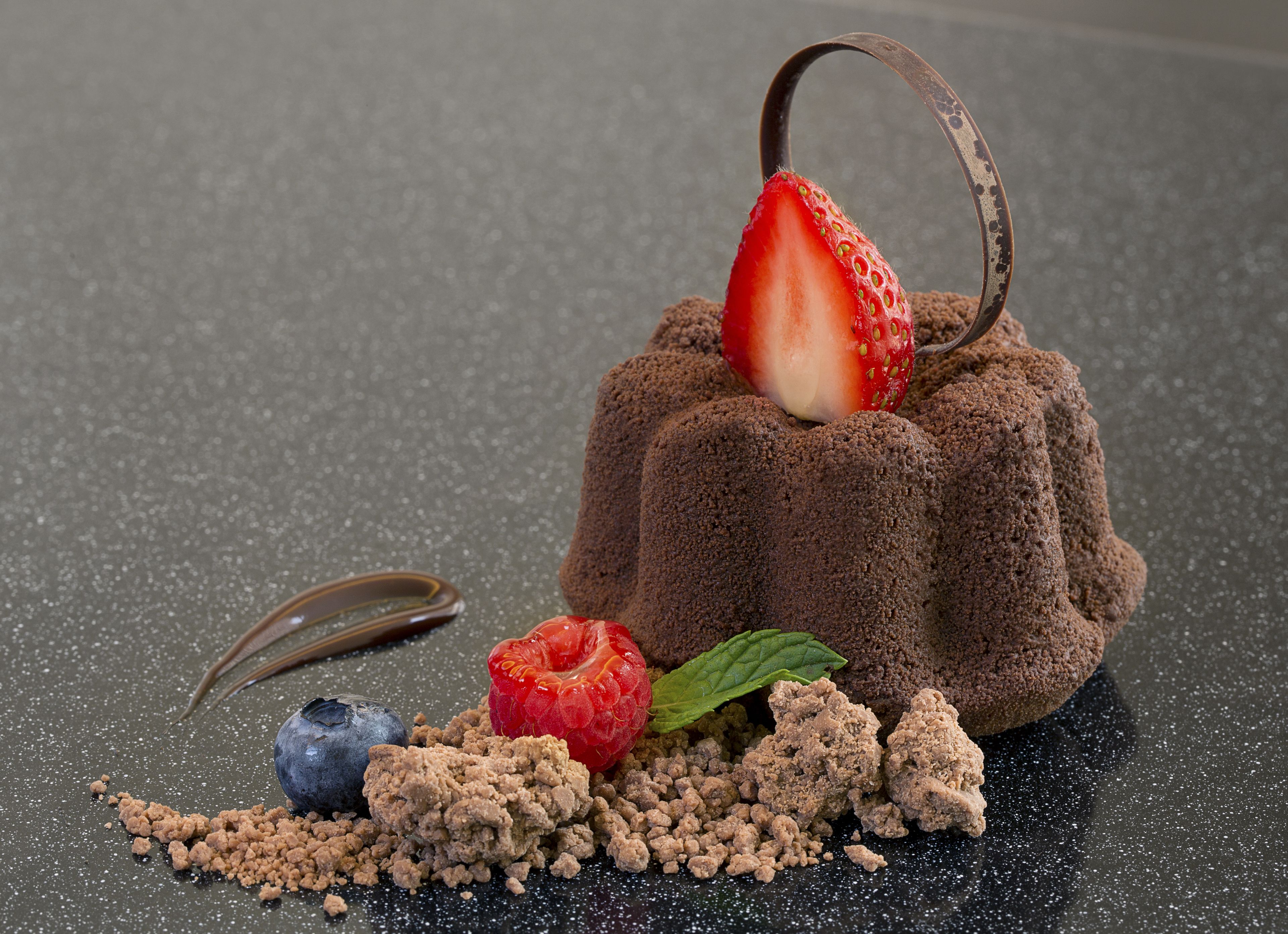 En chokladdessert med en jordgubbe ovanpå, omgiven av hallon, blåbär och smulor på en yta med en tydlig struktur.