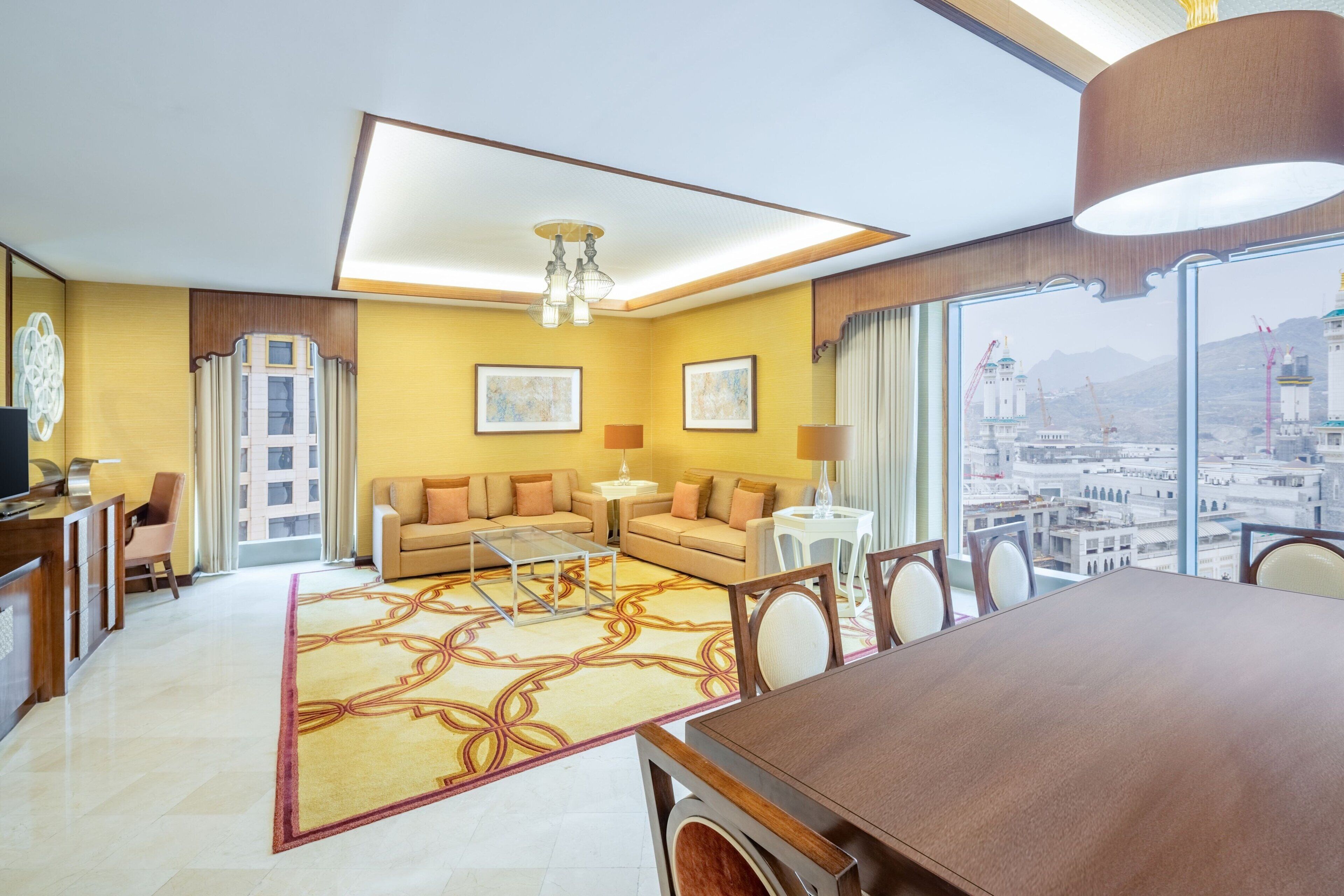 Une chambre d’hôtel dotée d’une grande table à manger, d’un ensemble de canapés, d’une table basse et offrant une vue sur le paysage urbain grâce à de grandes fenêtres.