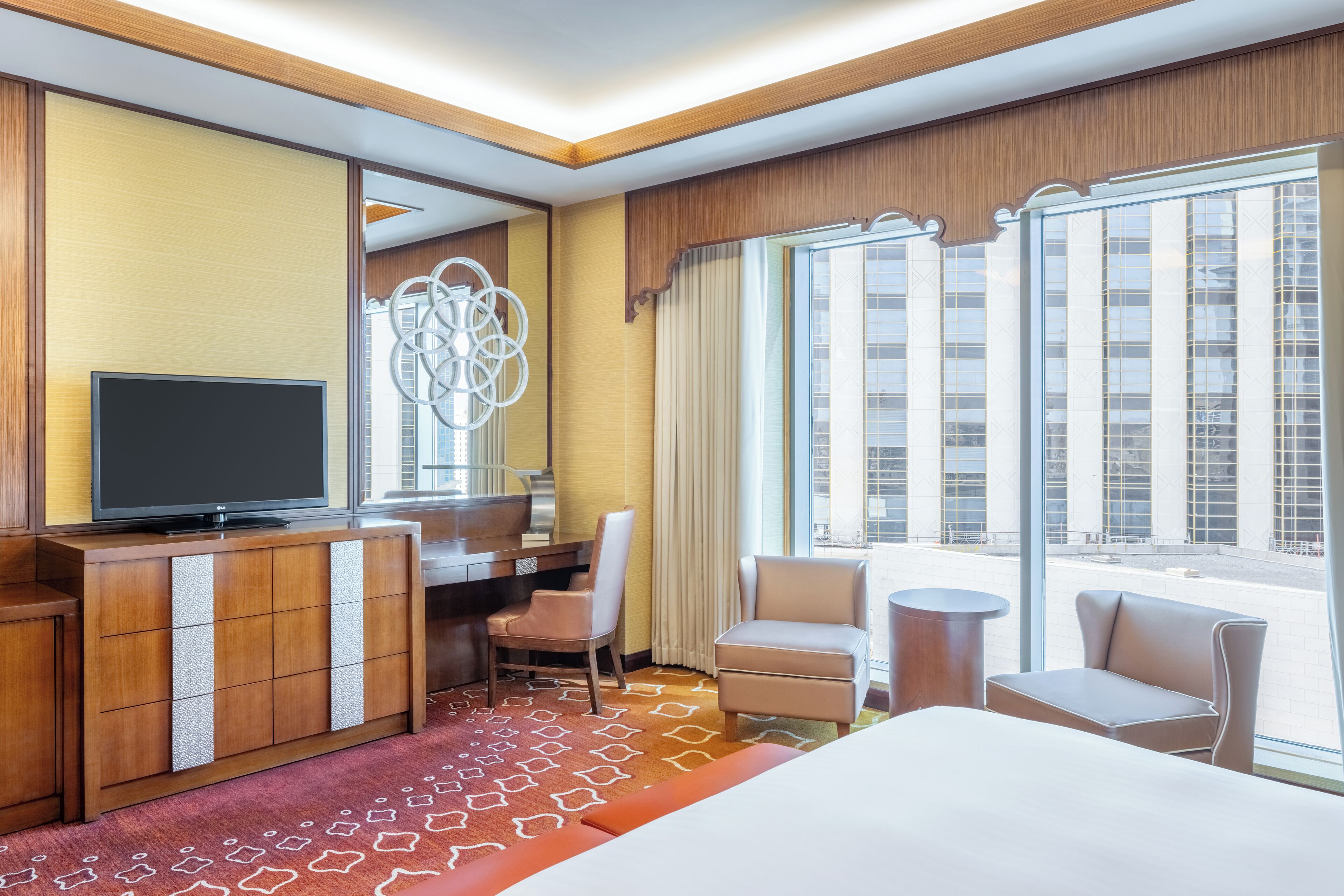A hotel room with a bed, a desk, a television, a chair, and a view of the city through large windows.