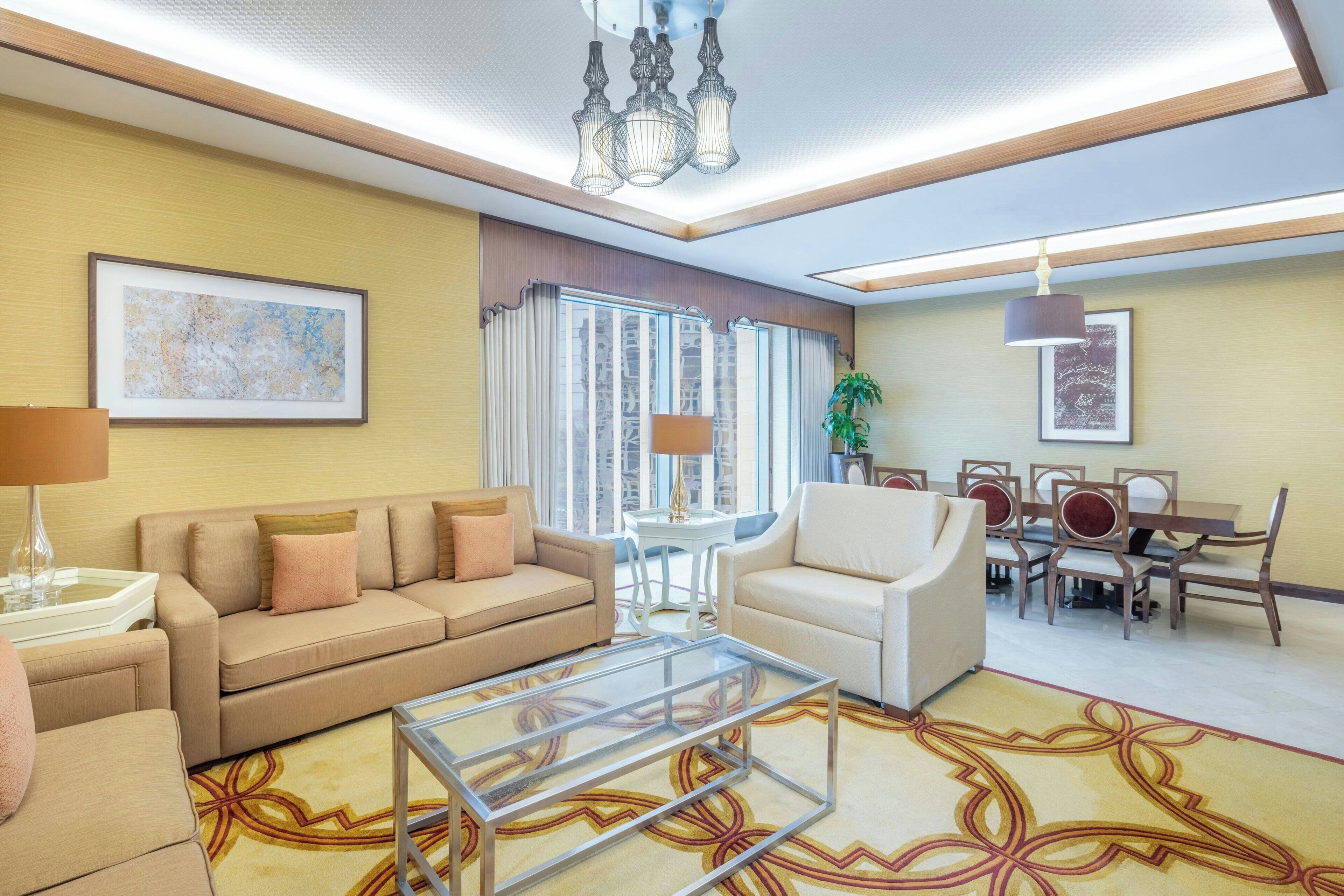 A modern hotel room with a sofa, armchair, dining table, and chairs. There is a large window with curtains, a chandelier, and a plant.