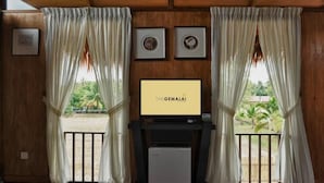 Quarto com paredes de madeira, duas janelas com cortinas brancas, uma televisão sobre um suporte e um frigorífico pequeno.