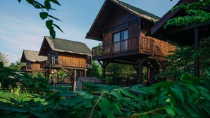 Uma cabana de madeira com telhado de palha, erguida sobre estacas, rodeada por vegetação exuberante.