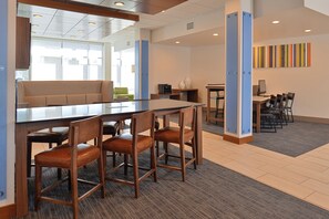 A modern hotel lobby with a long communal table, several wooden chairs, a sofa, and a small seating area with a green chair. - Holiday Inn Express & Suites Farmington Hills - Detroit by IHG (Farmington Hills)