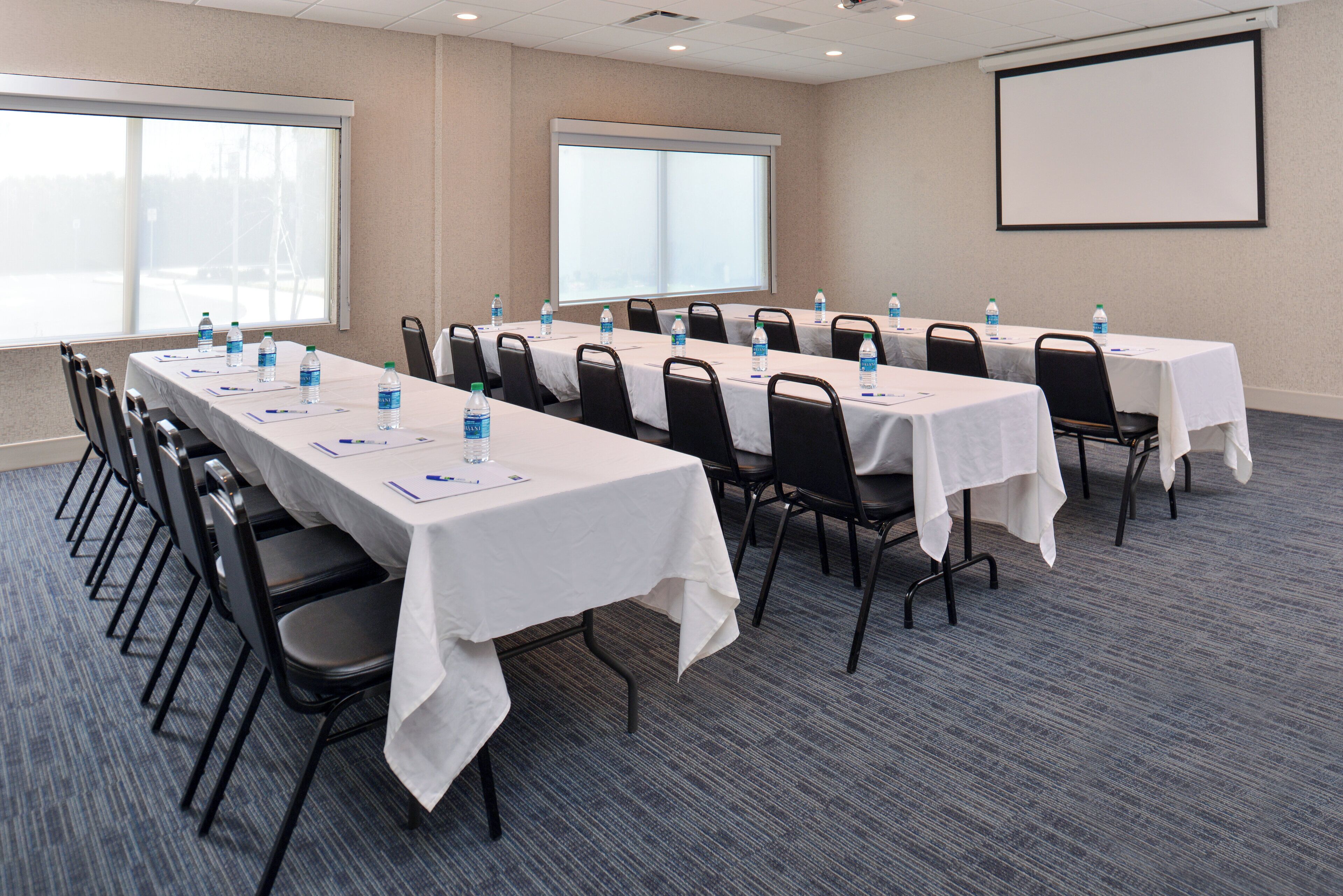 A conference room with long tables, chairs, water bottles, and a projector screen.