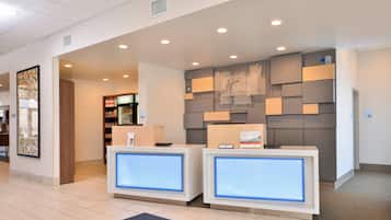 A hotel reception area with two reception desks, a wall-mounted information board, and a market area in the background.