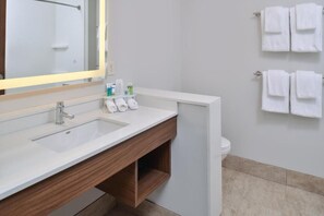 Een moderne badkamer met een witte wastafel, houten wastafelmeubel en een spiegel met verlichting. Er hangen witte handdoeken aan een rek en er is een toilet op de achtergrond te zien.