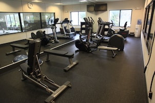 Un gimnasio con diferentes máquinas de ejercicio, ventanas amplias y un reloj en la pared.