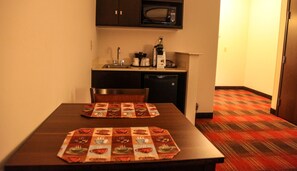 A small kitchenette with a microwave, coffee maker, and sink. There is a table with a placemat and a chair. The floor has a patterned carpet. - Holiday Inn Express & Suites Cumberland - La Vale by IHG (Cumberland)