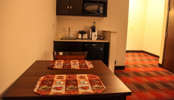 Una pequeña cocina equipada con microondas, cafetera y lavamanos. Hay una mesa con mantel individual y una silla. El piso tiene una alfombra estampada.