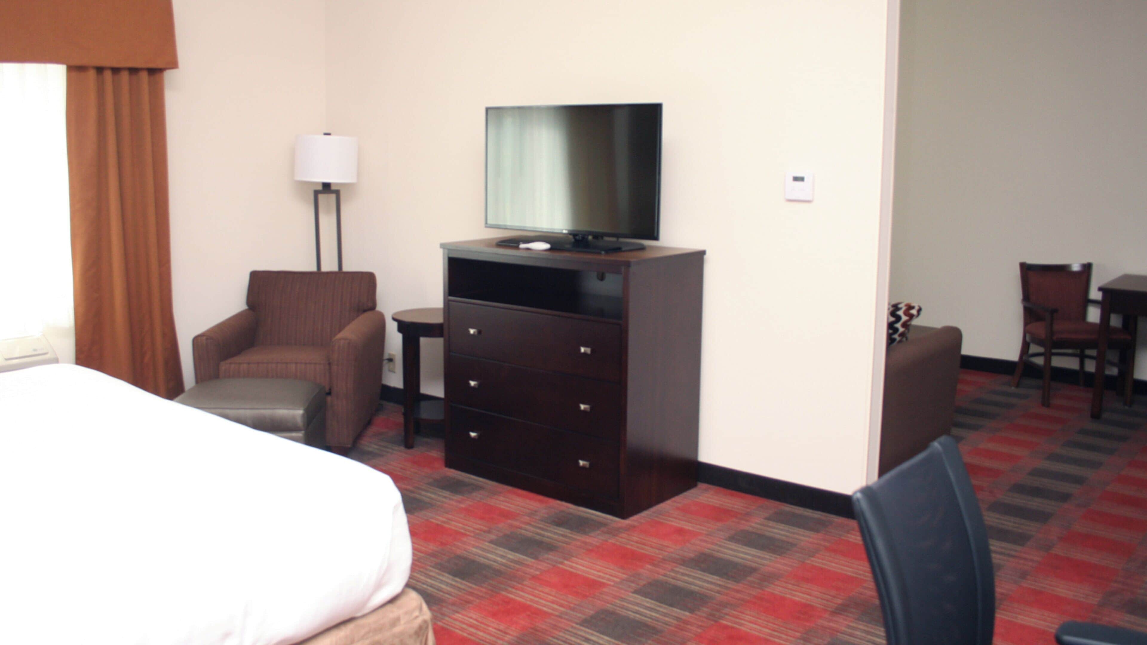 Habitación de hotel con cama, cómoda con televisor, una silla, escritorio y lámpara.