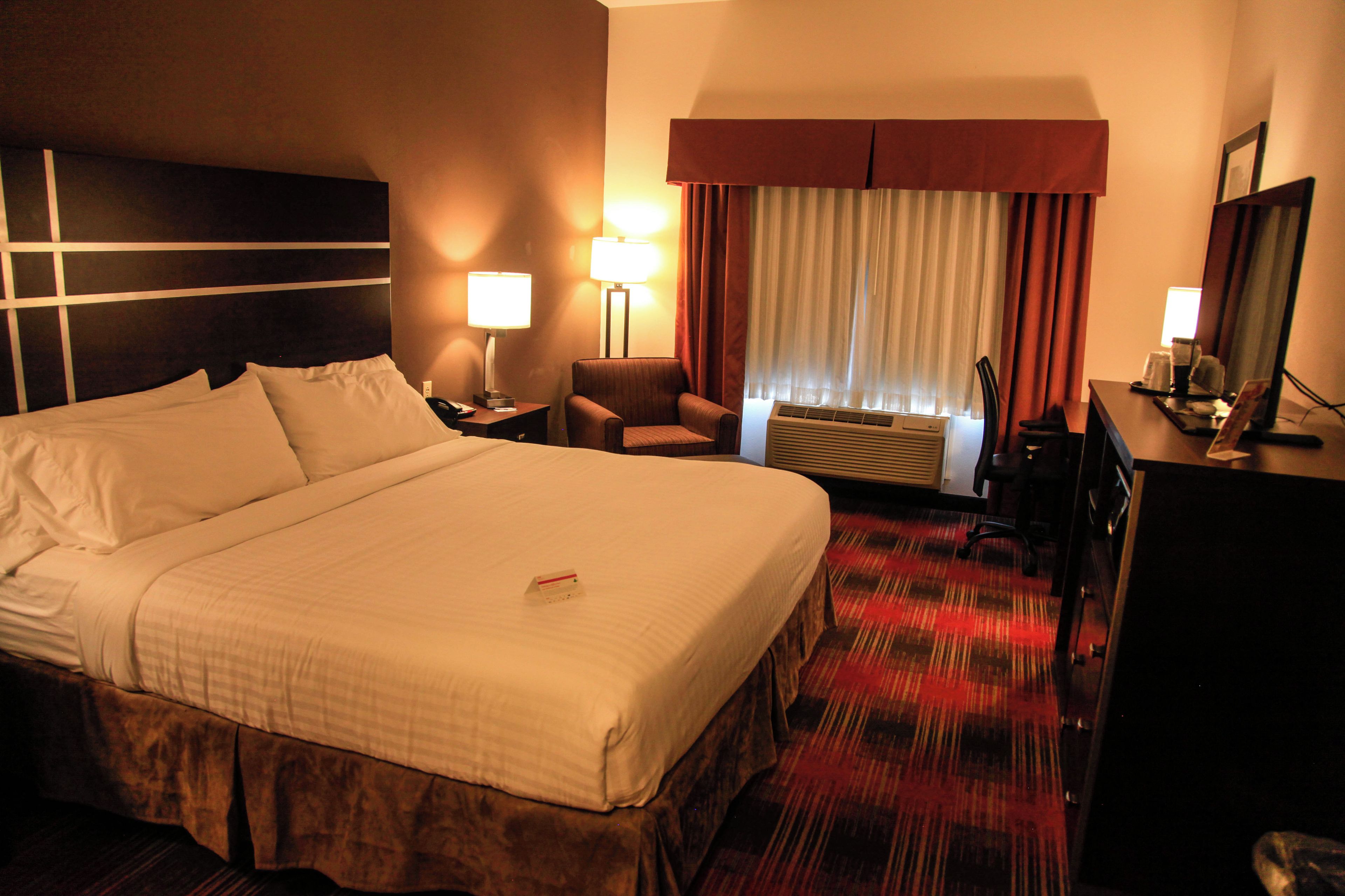 A hotel room with a large bed, two bedside tables, a chair, a desk, a lamp, an air conditioning unit, and a mirror.