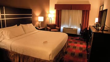 A hotel room with a large bed, two bedside tables, a chair, a desk, a lamp, an air conditioning unit, and a mirror.