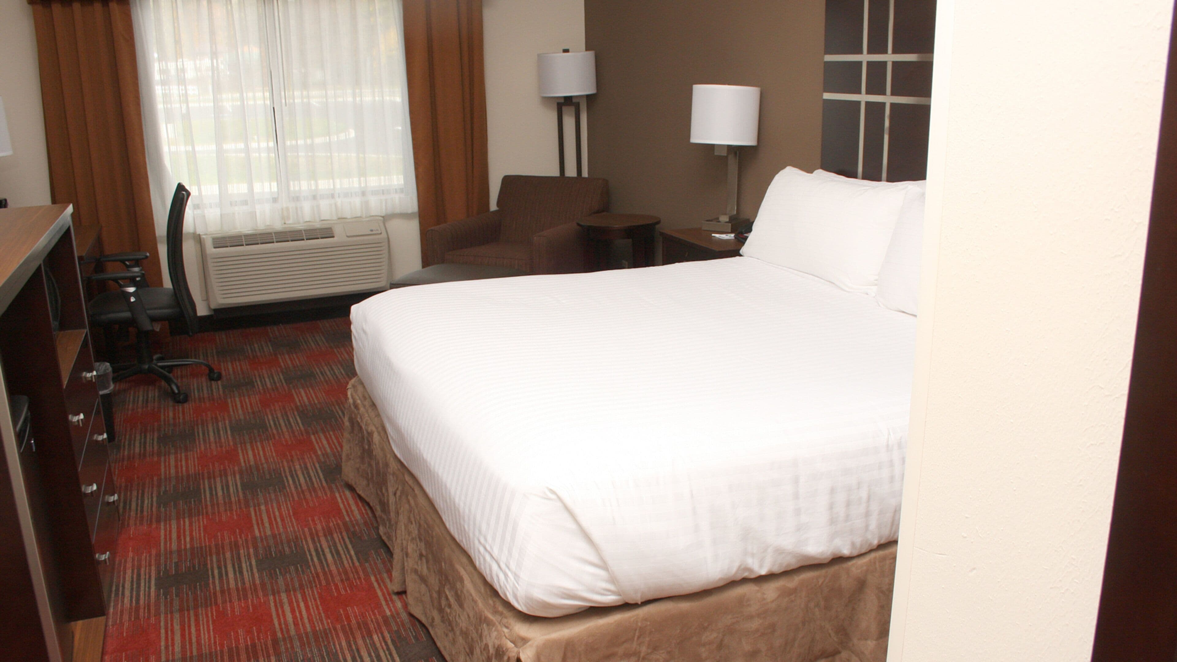 A hotel room with a bed, a desk, a chair, a lamp, and a window with curtains.