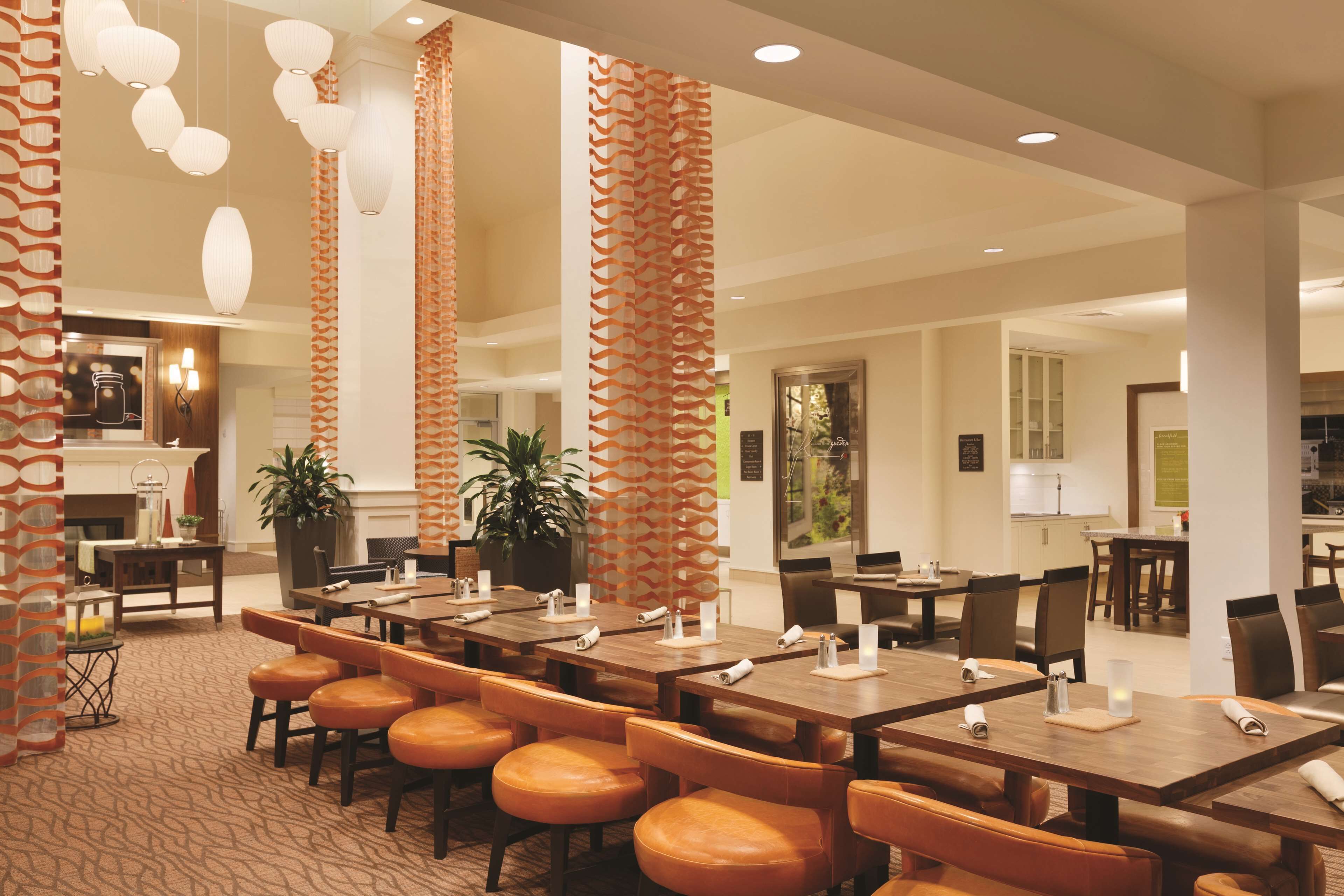 A hotel lobby with a long dining table, multiple chairs, and a kitchen area in the background.