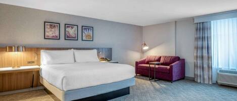 Habitación de hotel con cama, escritorio con lámpara, sofá rojo y ventana grande con cortinas.