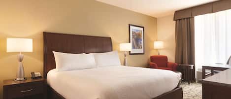 A hotel room with a large bed, two bedside tables with lamps, a red chair, a desk, and a framed picture on the wall.