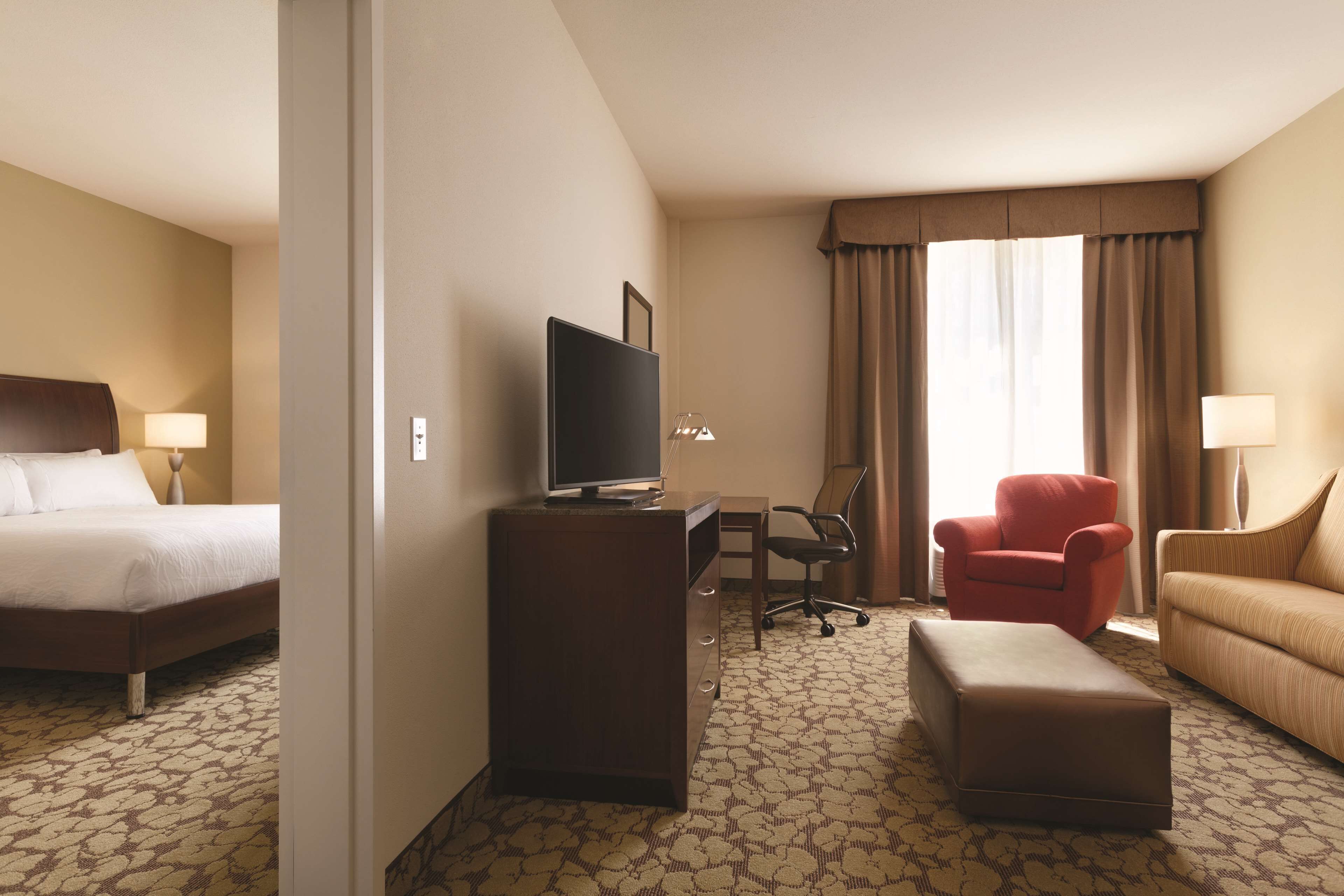 A hotel room with a bed, a desk with a television, a red armchair, a sofa, and patterned carpet.