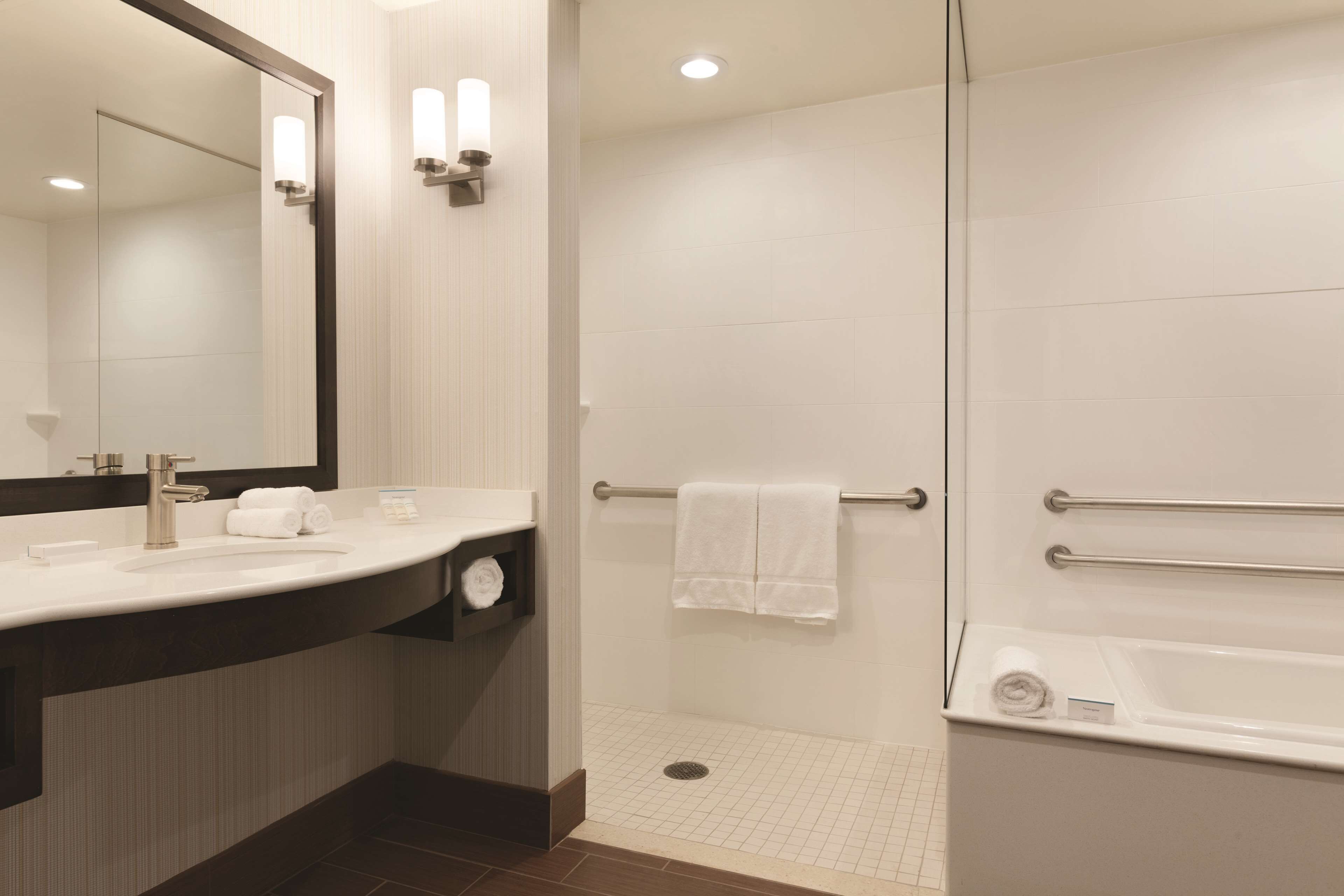 A modern bathroom with a large mirror, a sink with a towel holder, a shower with a grab bar, and a bathtub.