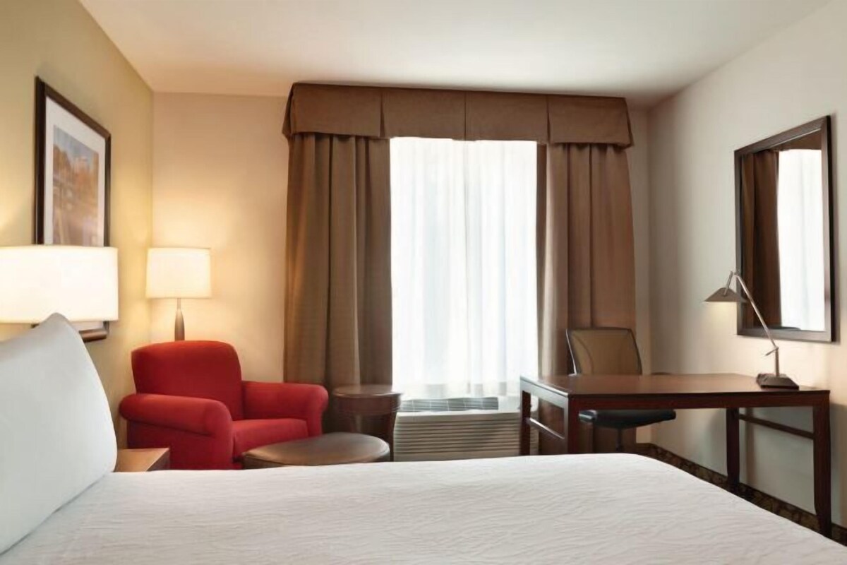 Ein Hotelzimmer mit einem Bett, einem Schreibtisch, einem Stuhl, einer Lampe und einem großen Fenster mit Vorhängen.