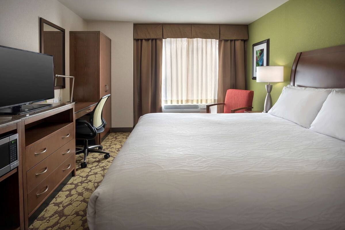 A hotel room with a bed, a desk with a computer, a chair, a television, and a window with curtains.