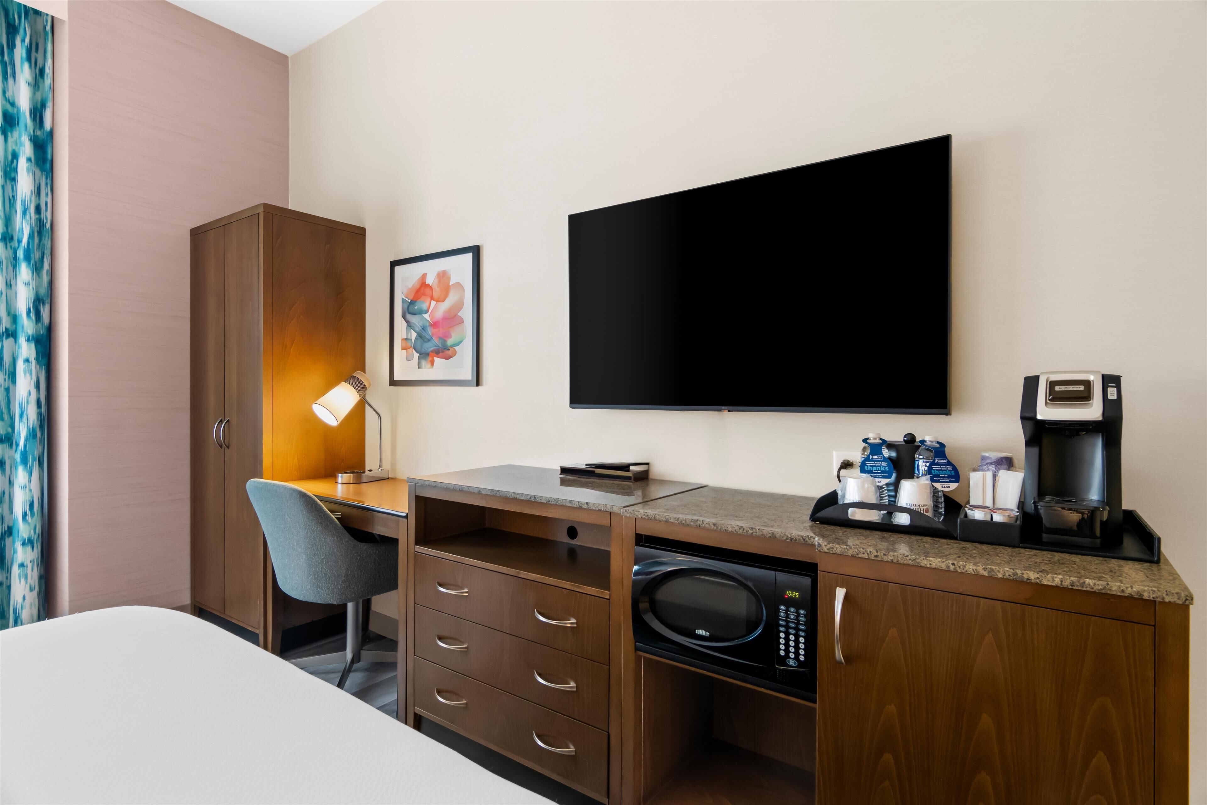 A hotel room with a bed, a desk with a lamp, a microwave, a coffee maker, and a flat-screen TV.