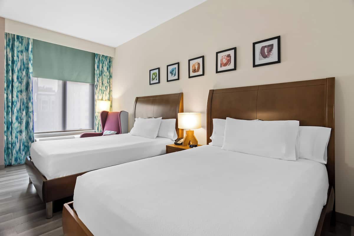 A hotel room with two beds, a window with curtains, a lamp, and framed pictures on the wall.
