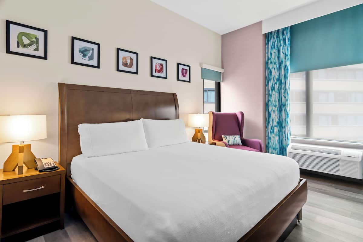 A hotel room with a large bed, bedside lamps, a chair, framed artwork on the wall, and an air conditioning unit.