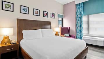 Ein Hotelzimmer mit einem großen Bett, Nachttischlampen, einem Sessel, gerahmten Bildern an der Wand und einer Klimaanlage.