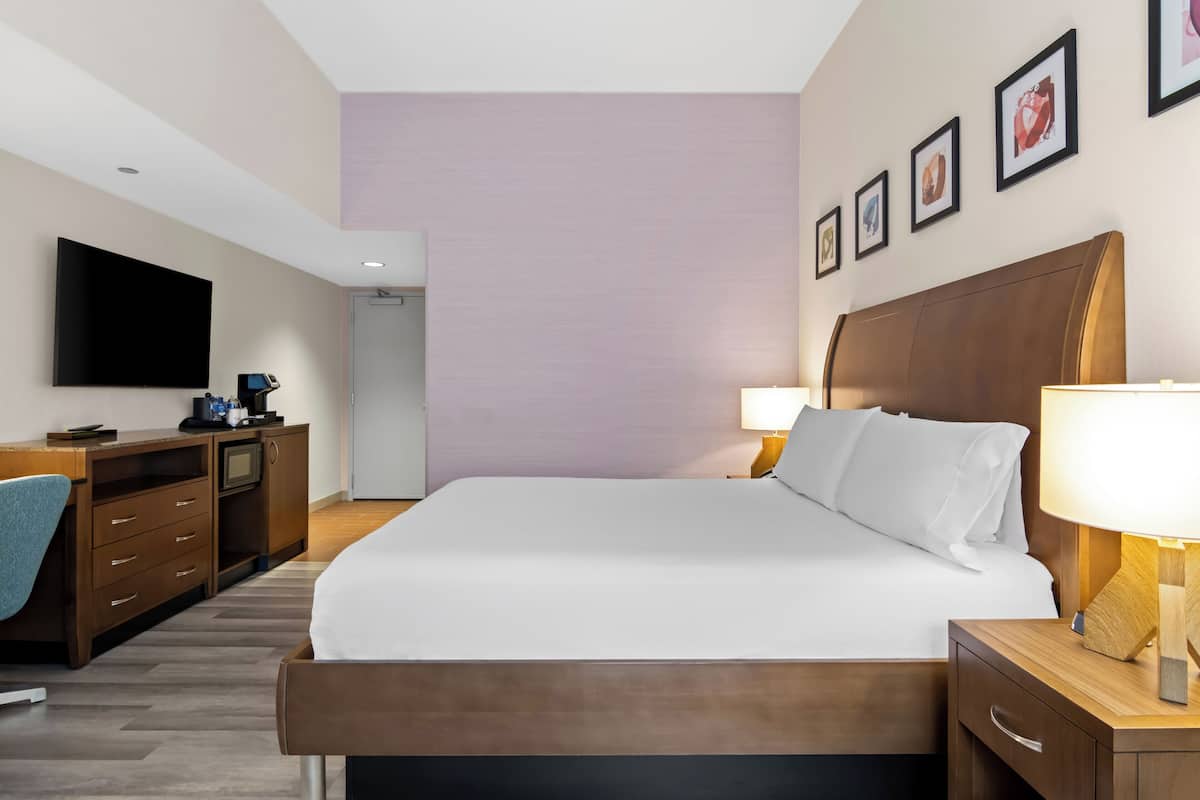 A hotel room with a bed, bedside table, lamp, desk with a computer, and a TV.